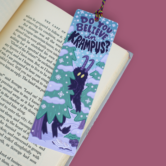 A decorative paper bookmark with a Krampus illustration and the text 'DO YOU BELIEVE IN KRAMPUS?' placed on an open book against a solid pink background.