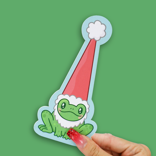 A die-cut sticker featuring a cute green frog wearing a red Santa hat with a white pom-pom and white faux-fur trim, held by a hand with a red glittery nail against a solid green background.