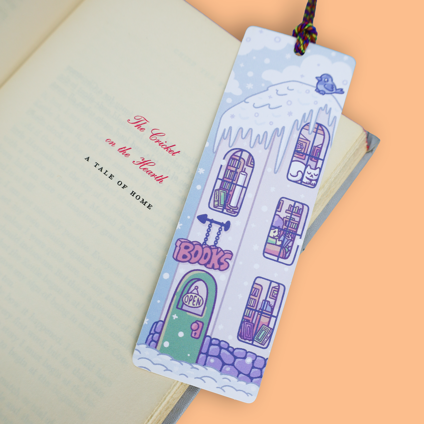 A decorative paper bookmark shaped like a snowy house with purple windows, a green door, and the word 'BOOKS' on the front, shown resting on an open book against a peach background.
