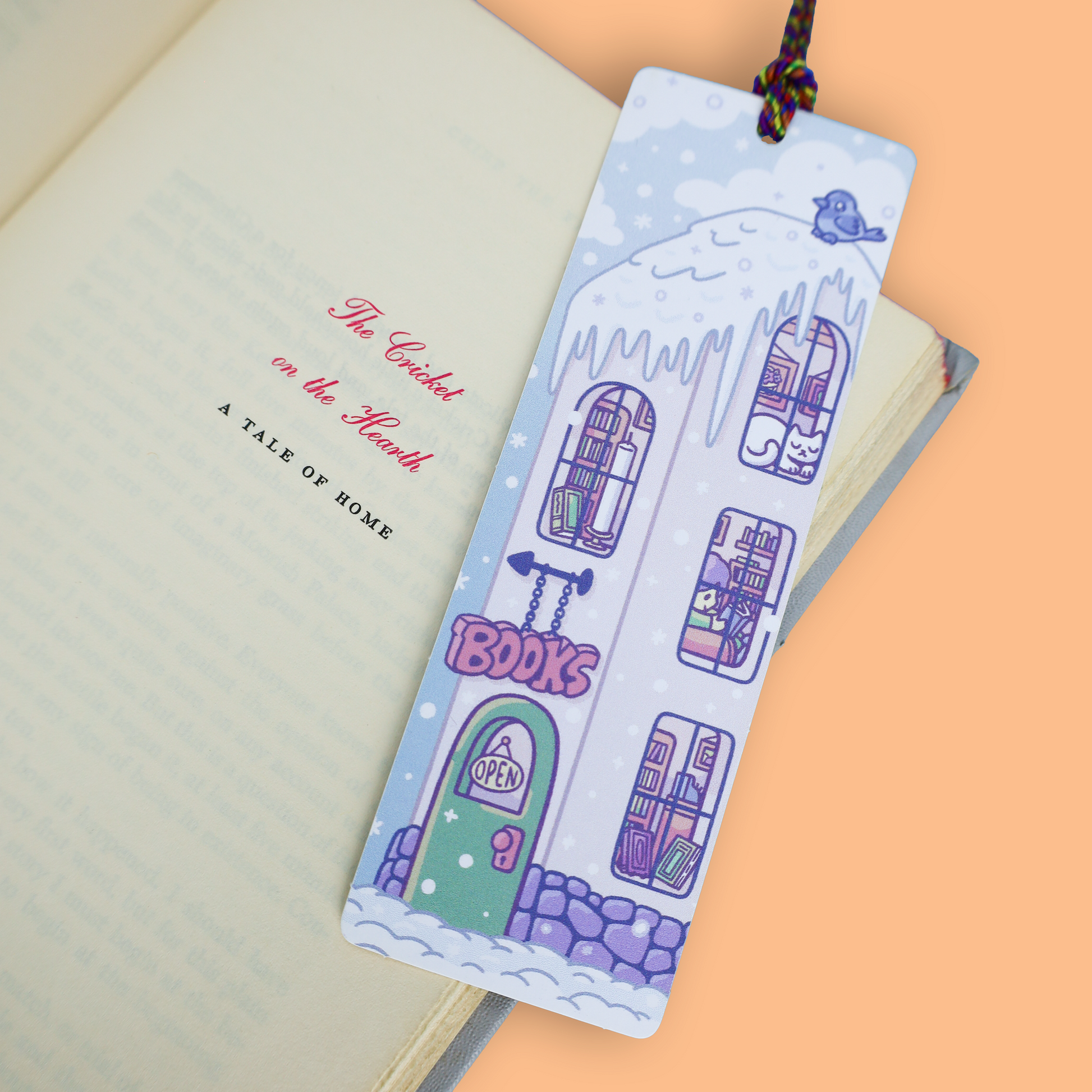 A decorative paper bookmark shaped like a snowy house with purple windows, a green door, and the word 'BOOKS' on the front, shown resting on an open book against a peach background.