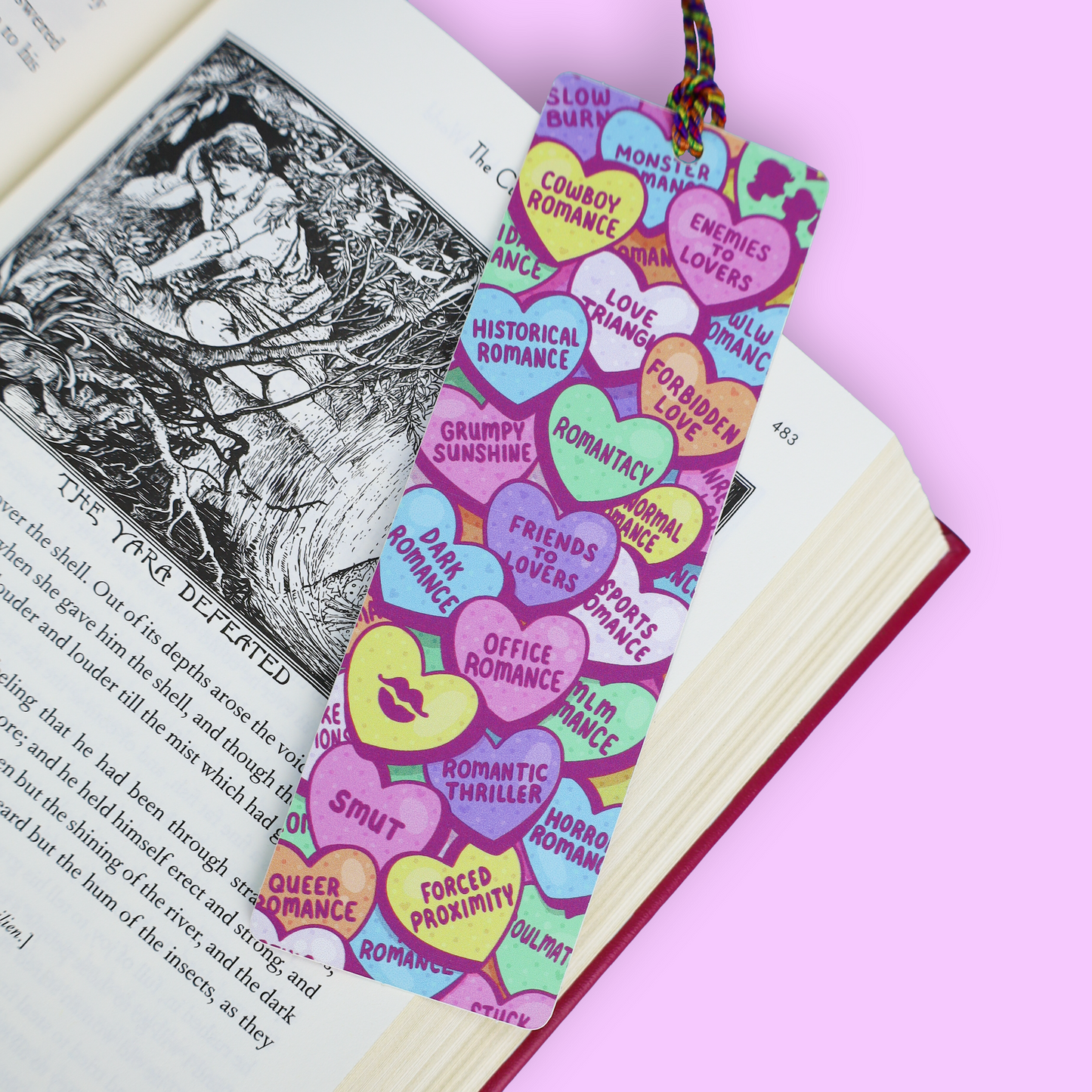 A colorful heart-shaped bookmark with candy-coated pastel sections and printed phrases like 'Romantic', 'Enemies Lovers', and 'Forbidden Love', placed on an open book against a pink background.