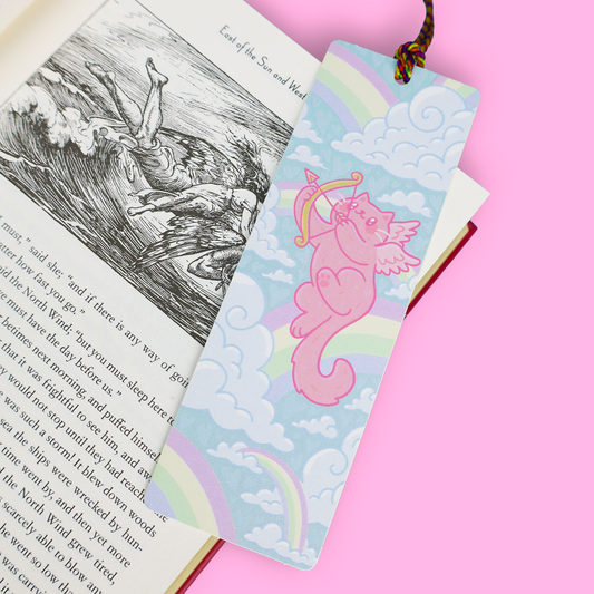 A decorative book bookmark with a pink cat holding a bow and arrow against a pastel rainbow and cloud background, shown resting on an open book over a solid pink surface.