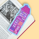 A colorful bookmark shaped like a pink house with a purple roof and the words 'Stories Book Shop' printed on it, placed on an open book page against a pale yellow background.