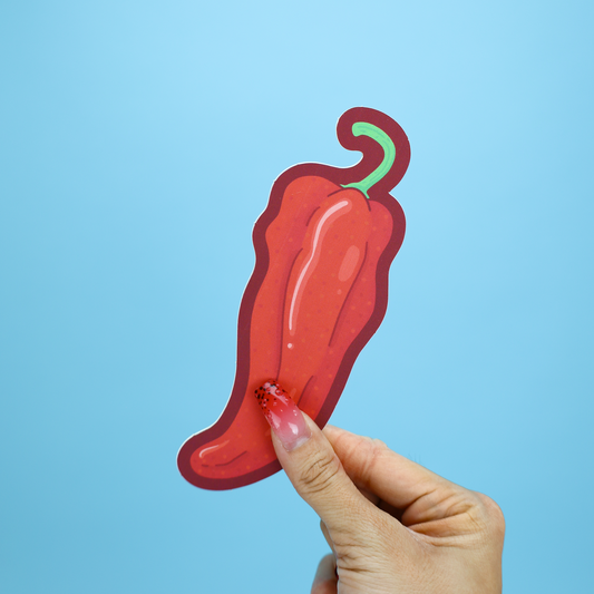 A hand with glittery pink nails holds a die-cut red chili pepper sticker or card against a solid light blue background.