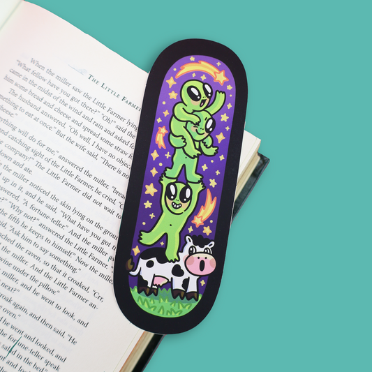 A black-bordered bookmark with a colorful cartoon illustration of green aliens stacking on a cow, placed on an open book against a teal background.
