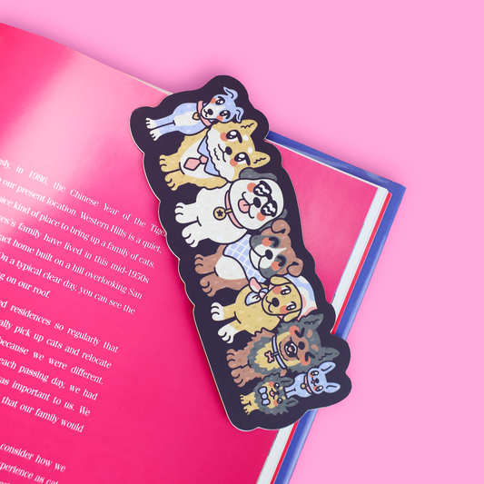 A die-cut sticker featuring a stacked arrangement of cute cartoon dogs in various breeds and colors, placed on top of an open pink book against a solid pink background.