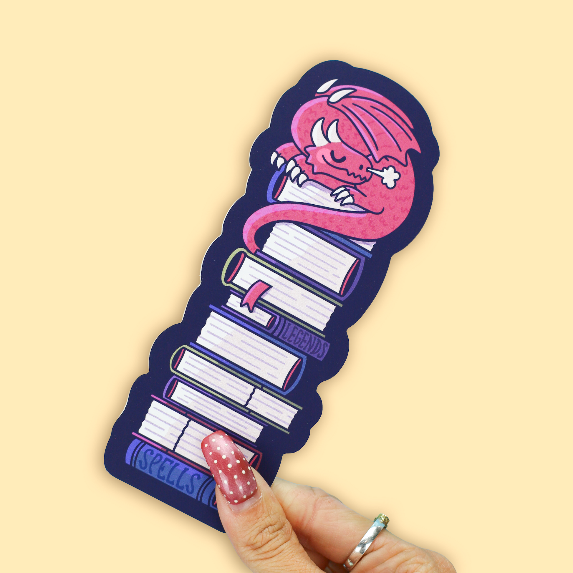 A hand with pink nail polish holds a die-cut sticker featuring a pink monster sleeping on top of a stack of books. The sticker has a dark navy blue border and the word 'ASPELLS' printed in blue at the bottom.