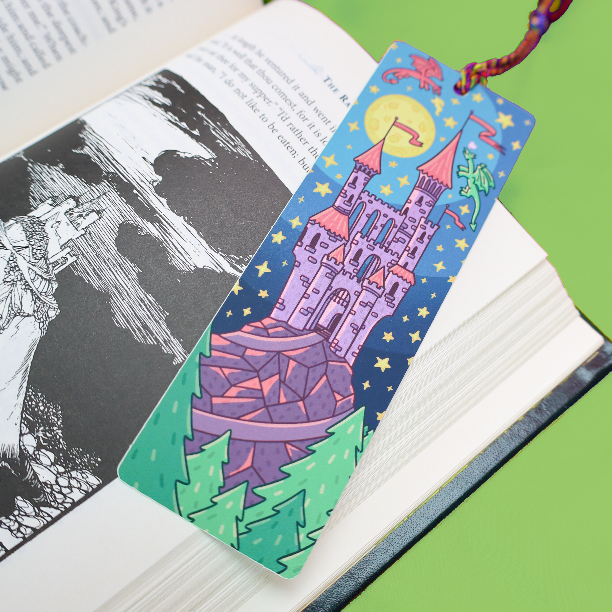 A decorative bookmark shaped like a castle in a fantasy night scene, resting on an open book. The bookmark features a purple castle under a yellow moon, green trees, and a red string loop at the top, photographed against a bright green background.