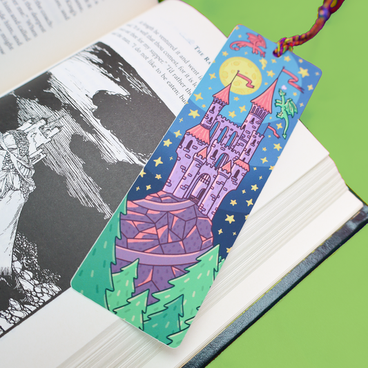 A decorative bookmark shaped like a castle in a fantasy night scene, resting on an open book. The bookmark features a purple castle under a yellow moon, green trees, and a red string loop at the top, photographed against a bright green background.