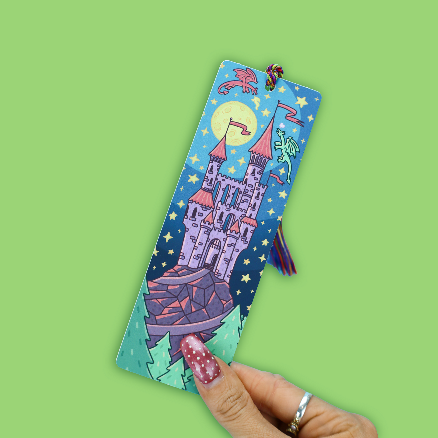 A hand with red glittery fingernails holds a decorative wooden bookmark featuring a colorful illustration of a castle under a full moon, set against a solid light green background.