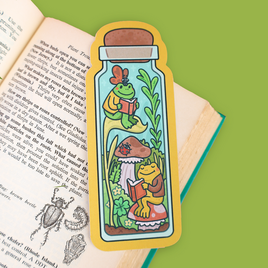 A die-cut sticker shaped like a glass bottle with cartoon frogs reading under a mushroom, placed on an open book against a bright green background.