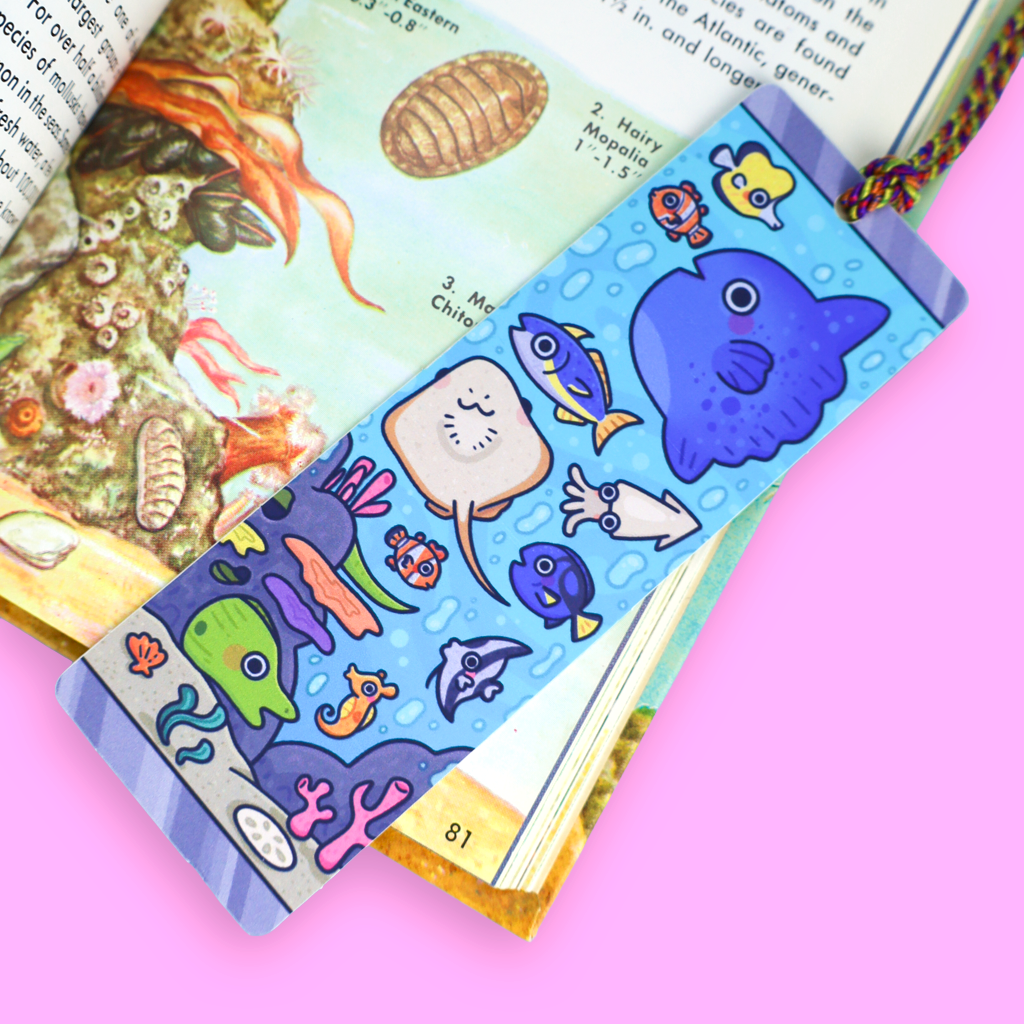 A colorful illustrated bookmark with cartoon sea creatures, including a blue whale, yellow fish, and coral, placed over an open children's book on a bright pink background.