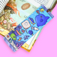 A colorful illustrated bookmark with cartoon sea creatures, including a blue whale, yellow fish, and coral, placed over an open children's book on a bright pink background.