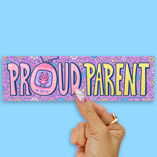 A hand holds a colorful rectangular sticker that reads 'PROUD PARENT' in playful outlined letters on a light blue background. The sticker has a pink and purple patterned base with hearts, stars, and a small character inside the letter O.