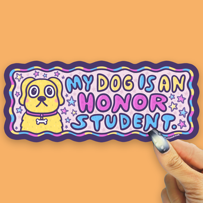 A die-cut sticker featuring a yellow cartoon dog and the text 'MY DOG IS AN HONOR STUDENT.' in colorful bubble letters, held by a hand against a solid orange background.