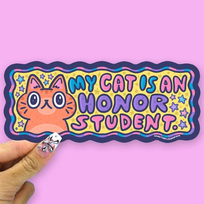 A hand holds a colorful die-cut sticker that reads 'MY CAT IS AN HONOR STUDENT.' against a solid pink background.