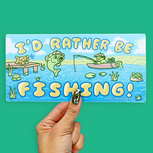A rectangular printed card or sticker with a colorful cartoon fishing scene and the text “I'D RATHER BE FISHING!” is held by a hand against a solid teal background. The card includes a small website credit at the bottom right.