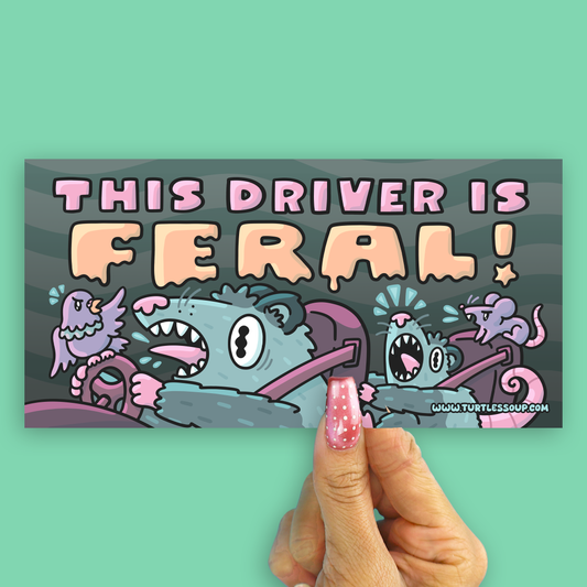 A hand with pink glitter nail polish holds a rectangular sticker against a solid mint green background. The sticker features a cartoon illustration of two gray monsters driving a car, with the text 'THIS DRIVER IS FERAL!' in pink and orange lettering.