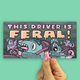 A hand with pink glitter nail polish holds a rectangular sticker against a solid mint green background. The sticker features a cartoon illustration of two gray monsters driving a car, with the text 'THIS DRIVER IS FERAL!' in pink and orange lettering.