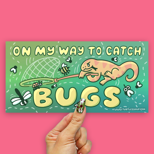 A rectangular printed card or sticker with a colorful cartoon design is held by a hand against a solid pink background. The design reads “ON MY WAY TO CATCH BUGS” with an illustrated orange cat catching bugs on a green-to-teal gradient background.