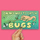 A rectangular printed card or sticker with a colorful cartoon design is held by a hand against a solid pink background. The design reads “ON MY WAY TO CATCH BUGS” with an illustrated orange cat catching bugs on a green-to-teal gradient background.