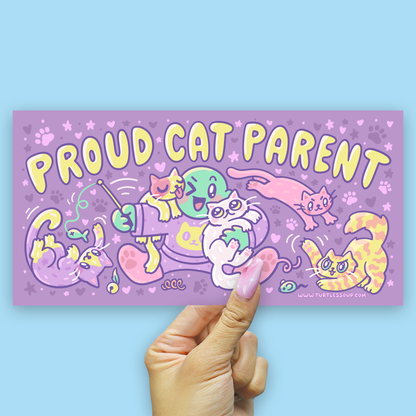 A hand with pink nail polish holds a rectangular purple sticker that reads 'PROUD CAT PARENT' in large yellow letters, surrounded by colorful cartoon cats and paw prints, against a light blue background.