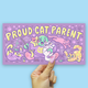 A hand with pink nail polish holds a rectangular purple sticker that reads 'PROUD CAT PARENT' in large yellow letters, surrounded by colorful cartoon cats and paw prints, against a light blue background.