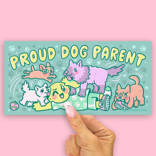 A rectangular sticker or card with a pastel illustration of playful dogs and the text 'PROUD DOG PARENT' is held by a person's hand against a solid pink background.