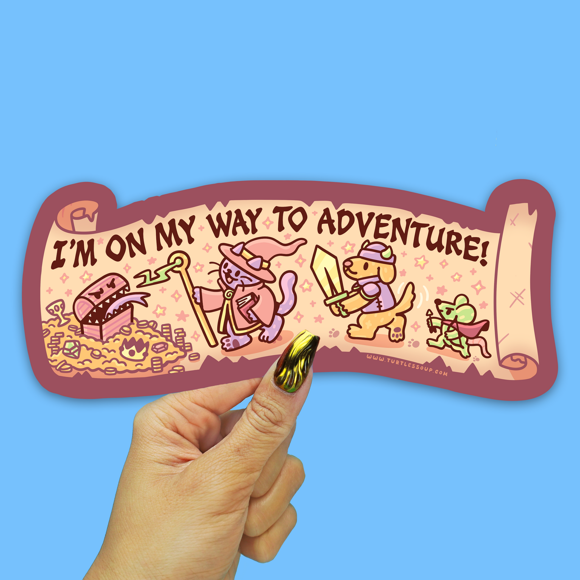 A hand with green glitter nail polish holds a die-cut sticker that reads 'I'M ON MY WAY TO ADVENTURE!' against a solid light blue background. The sticker features cartoon illustrations of a dragon, a wizard, a dog, and a frog on a parchment scroll design.