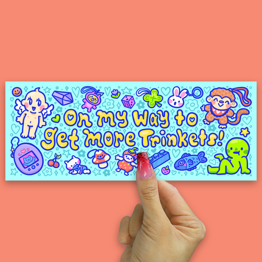 A rectangular sticker with a light blue background and colorful cartoon illustrations, held by a hand with long pink nails against a solid coral background. The sticker features the text 'Oh my way to Get more Trinkets!' in yellow and blue bubble letters, surrounded by cute cartoon characters and objects like a baby dragon, rabbit, monkey, and a toy phone.