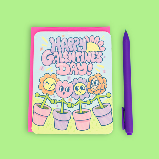 A square greeting card with a pink envelope, featuring a colorful 'Happy Galentine's Day!' design with cartoon flowers in pots, shown next to a purple pen on a light green background.