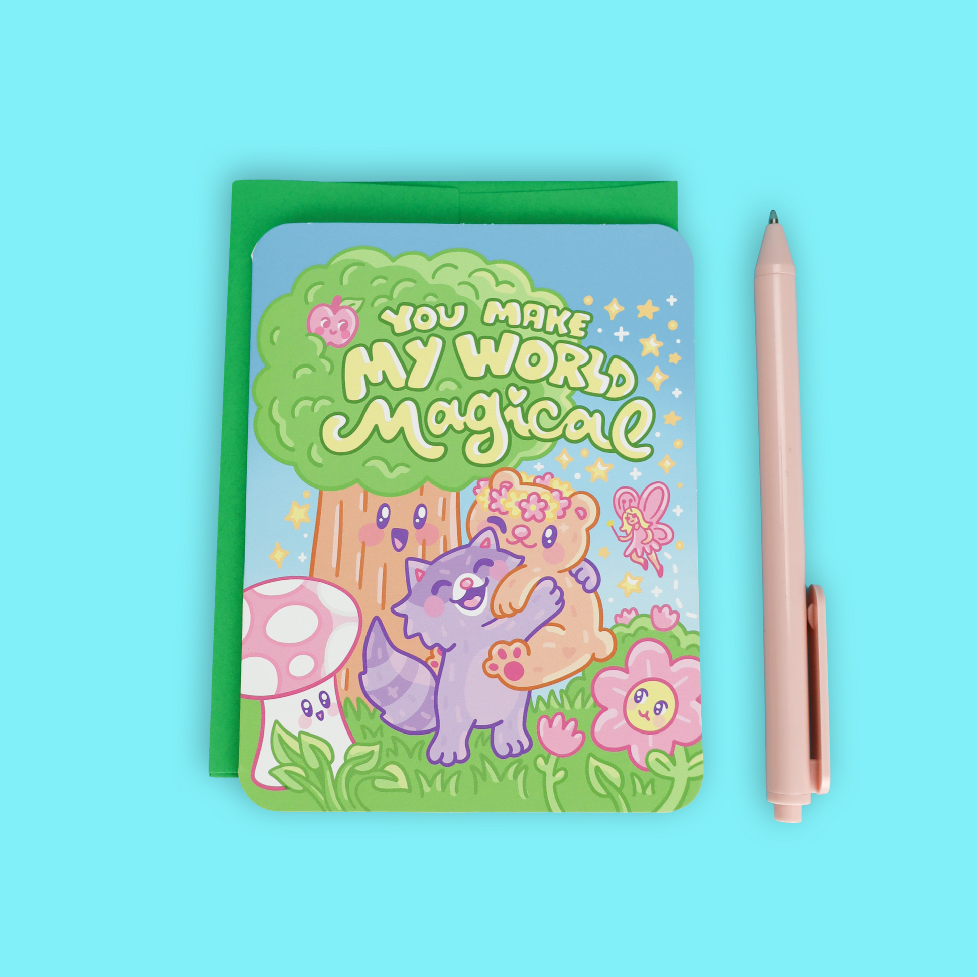 A pastel pink pen placed next to a small illustrated notebook or card with a green backing. The front features a whimsical forest illustration with the text 'YOU MAKE MY WORLD Magical,' a purple cat, a teddy bear, flowers, and a butterfly, all on a light blue background.