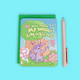 A pastel pink pen placed next to a small illustrated notebook or card with a green backing. The front features a whimsical forest illustration with the text 'YOU MAKE MY WORLD Magical,' a purple cat, a teddy bear, flowers, and a butterfly, all on a light blue background.