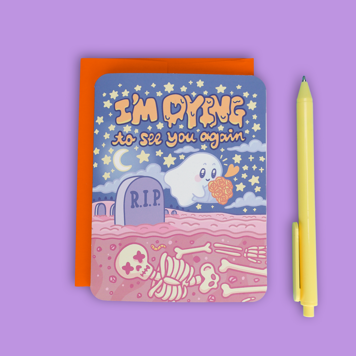 A square greeting card with a Halloween-themed illustration reading 'I'm DYING to see you again,' featuring a ghost, stars, a tombstone, and skeleton bones, shown with an orange backing card and a yellow pen on a lavender background.