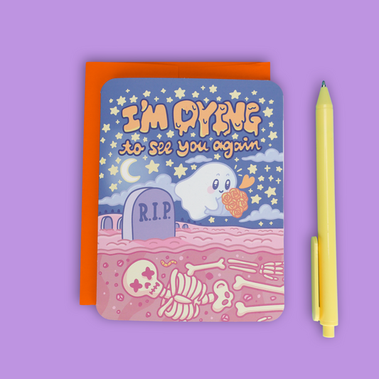 A square greeting card with a Halloween-themed illustration reading 'I'm DYING to see you again,' featuring a ghost, stars, a tombstone, and skeleton bones, shown with an orange backing card and a yellow pen on a lavender background.