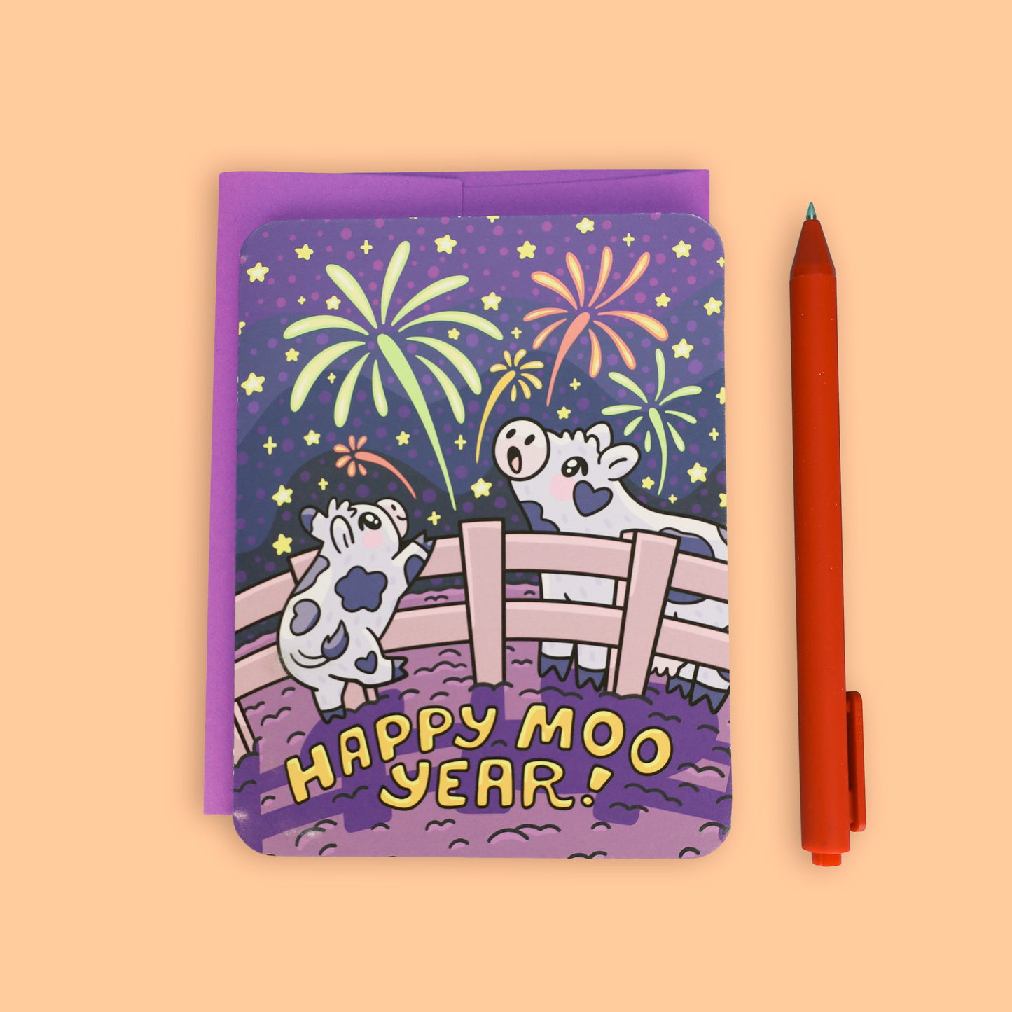 A purple greeting card with a cartoon cow and horse under fireworks and the text 'HAPPY MOO YEAR!' shown on a peach background next to an orange pen.