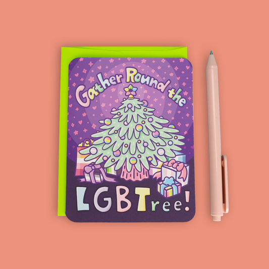A square greeting card with rounded corners featuring a colorful illustration of a Christmas tree and the text 'Gather Round the LGBTree!' in pastel letters, shown on a peach background next to a lime green envelope and a light pink pen.