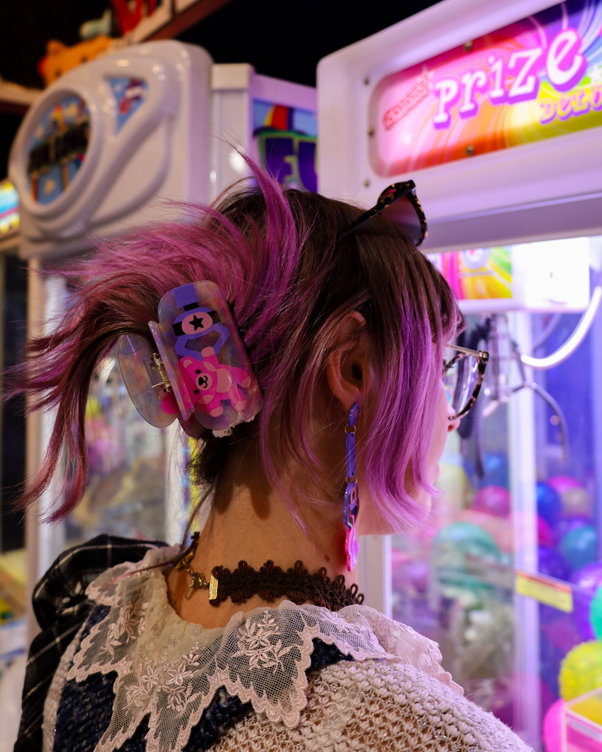 A person wearing a pink and purple claw machine cartoon bear hair clip in a brightly lit arcade room with claw machines.