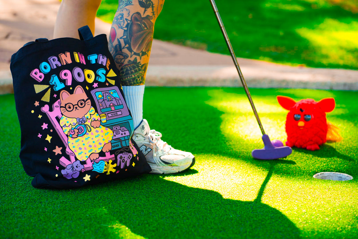 A tote bag with a colorful cartoon graphic and the text 'BORN IN THE 1900s' sits on bright green artificial turf next to a person's tattooed legs holding a purple putter. A red toy is also visible in the background.