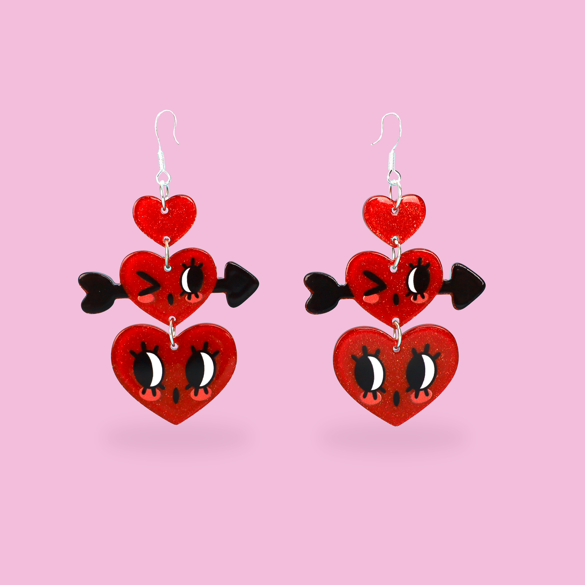 A pair of dangling heart earrings on a solid pink background. Each earring features three stacked red heart charms with black and white cartoon eyes and a small black heart-shaped arrowhead on the side.