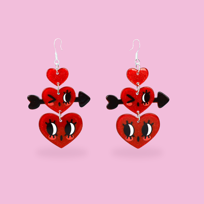 A pair of dangling heart earrings on a solid pink background. Each earring features three stacked red heart charms with black and white cartoon eyes and a small black heart-shaped arrowhead on the side.