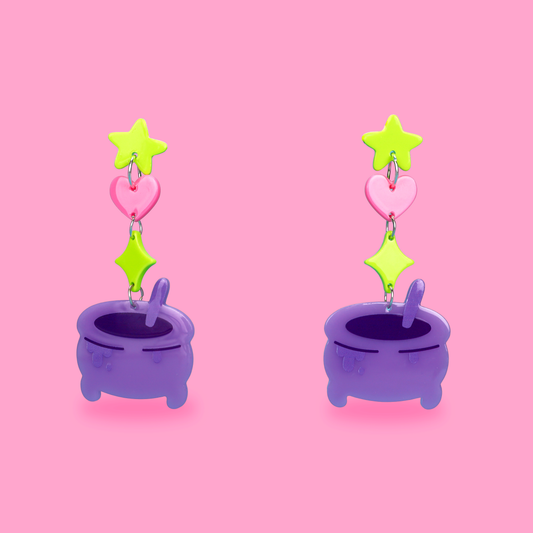 A pair of whimsical triple-drop dangle earrings featuring lime green star and diamond charms, a pink heart charm, and a purple cauldron pendant, set against a solid pink background.