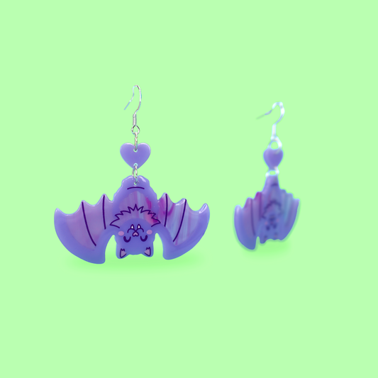 A pair of dangling bat-shaped earrings in glossy purple and lavender, shown on a solid light green background.