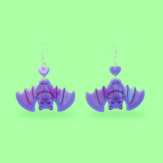A pair of dangling bat-shaped earrings in glossy purple and lavender, shown on a solid light green background.