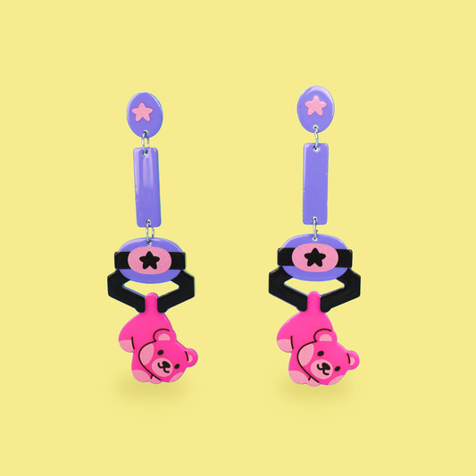 A pair of dangling earrings on a pale yellow background. Each earring has a lavender oval stud with a pink star, a lavender rectangular drop, a black and pink circular section with a star, and a dangling pink bear charm with black facial features.