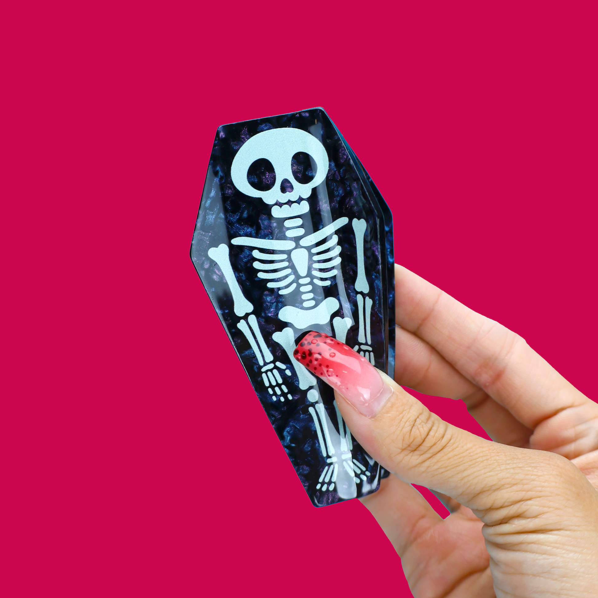 A hand with red glittery nails holds a small coffin-shaped decorative object featuring a white skeleton print on a dark blue marbled background, set against a solid bright pink background.