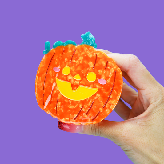 A hand holding a small orange jack-o'-lantern-shaped item with a yellow smiley face and blue stem details against a solid purple background.