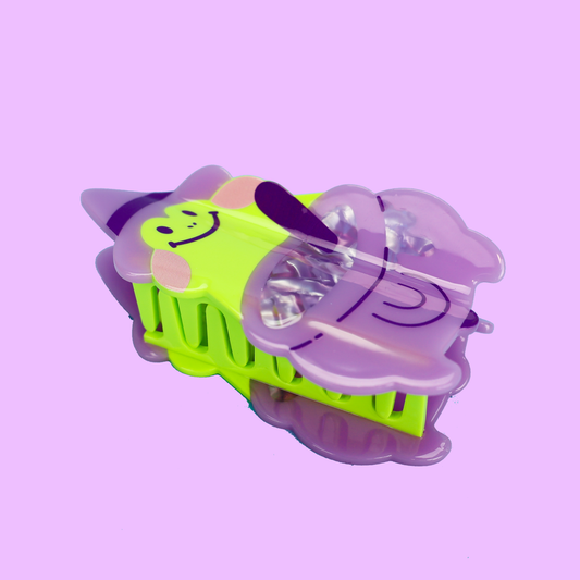 A hand with red glittery fingernails holds a small novelty toy shaped like a wizard hat and pot. The toy is translucent purple with a bright neon yellow-green face and black smiley details, set against a solid light purple background.