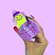 A hand with red glittery fingernails holds a small novelty toy shaped like a wizard hat and pot. The toy is translucent purple with a bright neon yellow-green face and black smiley details, set against a solid light purple background.