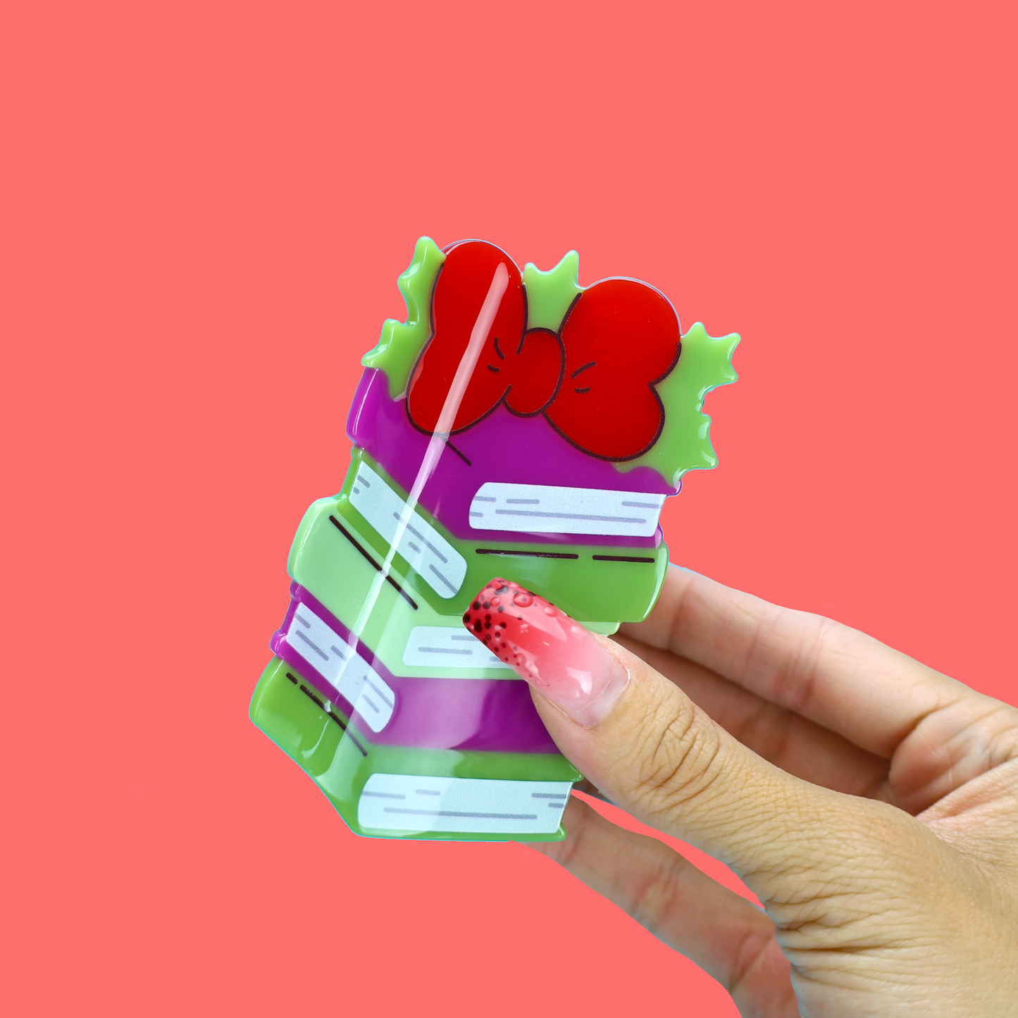 A hand with pink glitter nail polish holds a small, colorful, gift-box-shaped accessory against a solid coral pink background.